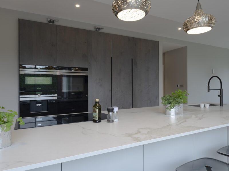 Mirrored cabinet back, with grey flat cupboards that go up to the ceiling with build in ovens, storage shelves, and an island with a marble