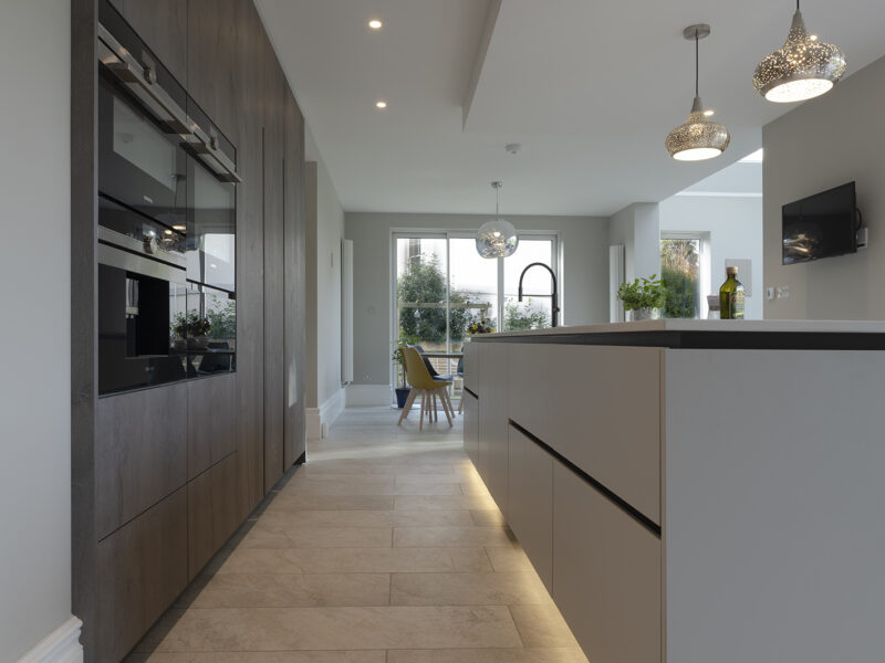 Mirrored cabinet back, with grey flat cupboards that go up to the ceiling with build in ovens, storage shelves, and an island with a marble