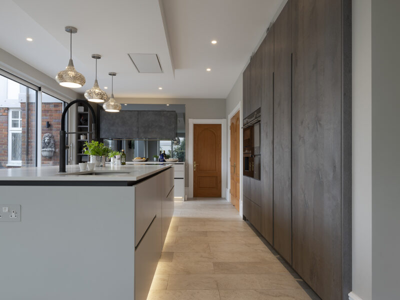 Mirrored cabinet back, with grey flat cupboards that go up to the ceiling with build in ovens, storage shelves, and an island with a marble