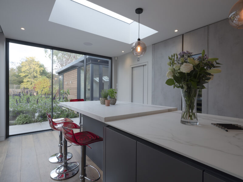 Mirrored cabinet back, with grey flat cupboards that go up to the ceiling with build in ovens, storage shelves, and an island with a marble