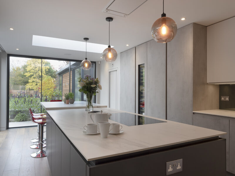 Mirrored cabinet back, with grey flat cupboards that go up to the ceiling with build in ovens, storage shelves, and an island with a marble