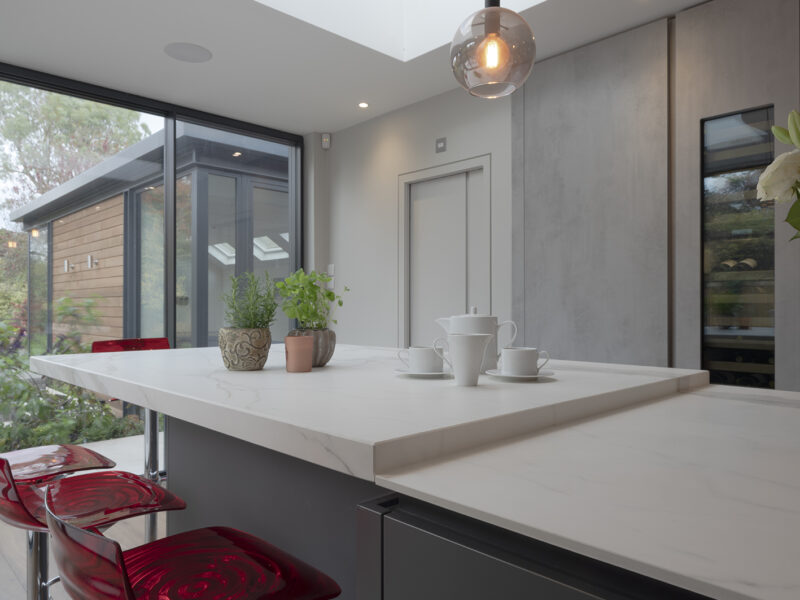 Mirrored cabinet back, with grey flat cupboards that go up to the ceiling with build in ovens, storage shelves, and an island with a marble