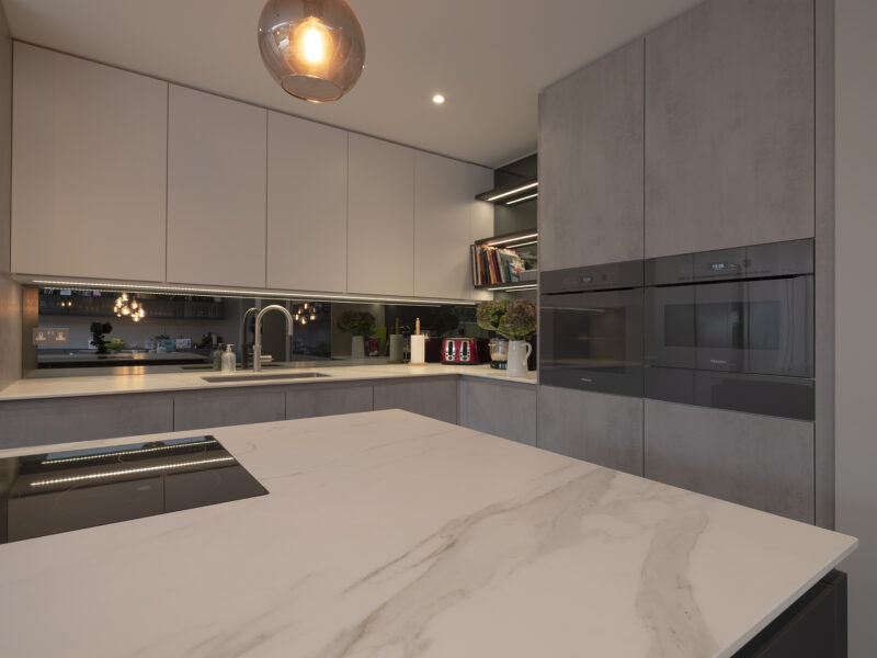 Mirrored cabinet back, with grey flat cupboards that go up to the ceiling with build in ovens, storage shelves, and an island with a marble